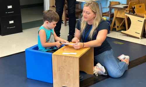 Occupational therapist Bianca sits on the floor next to Kolton who is cutting a piece of paper with scissors sitting in a blue chair.
