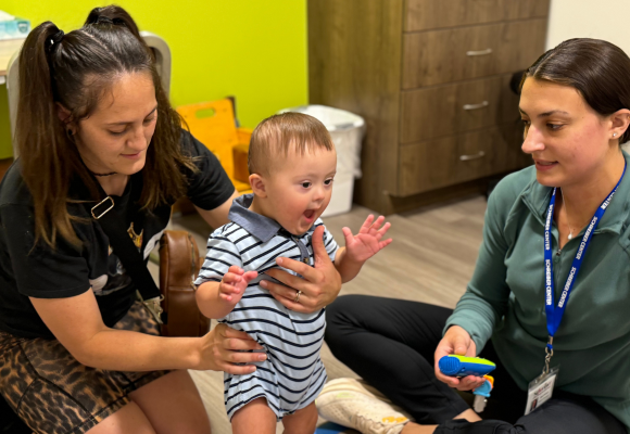 Kyrain standing with his mom, Tyara, supporting him with physical therapist Alexa sitting nearby.