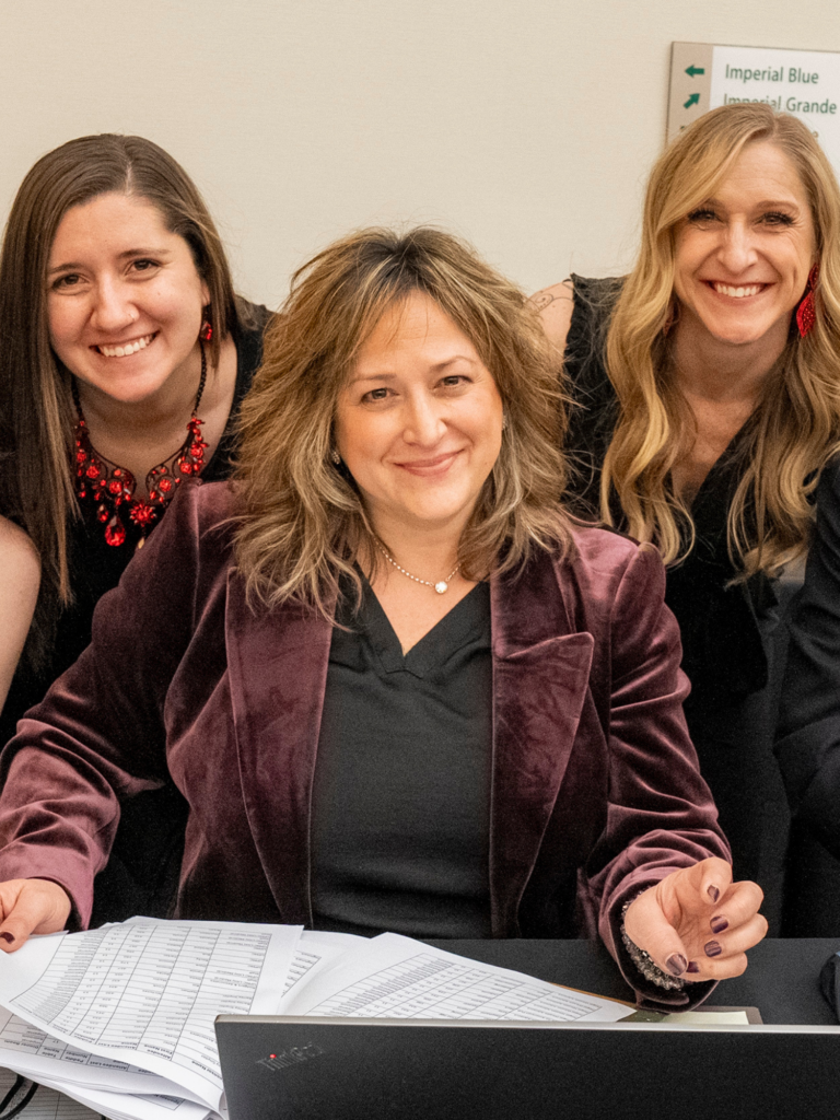 Three adults smiling while the adult in the middle sits at a desk and the other two stand behind them.