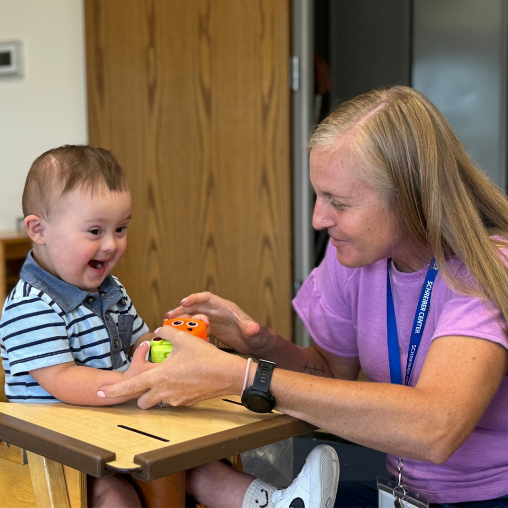 A child in a highchair smiling and holding items in their hands with a therapist assisting them.