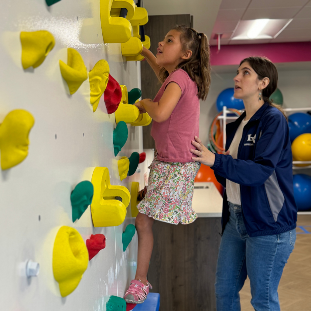 A child climbing on a colorful rock wall with a therapist guiding them.