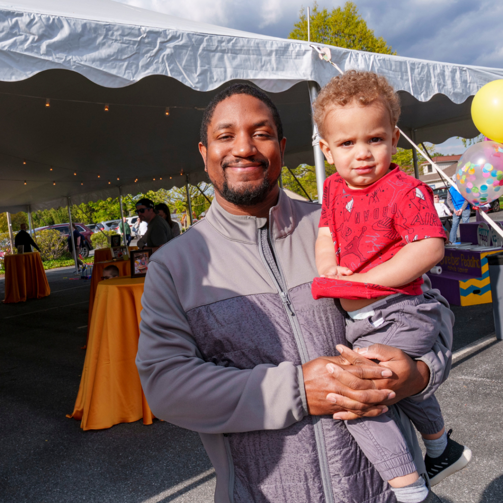 A Schreiber Family of two, an adult holding a child, both smiling in front of a white event tent.
