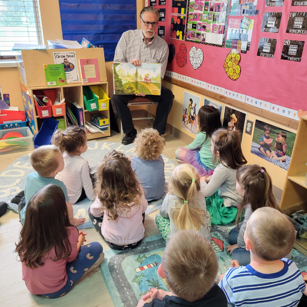 middle aged man reading picture book to a room full of preschoolers