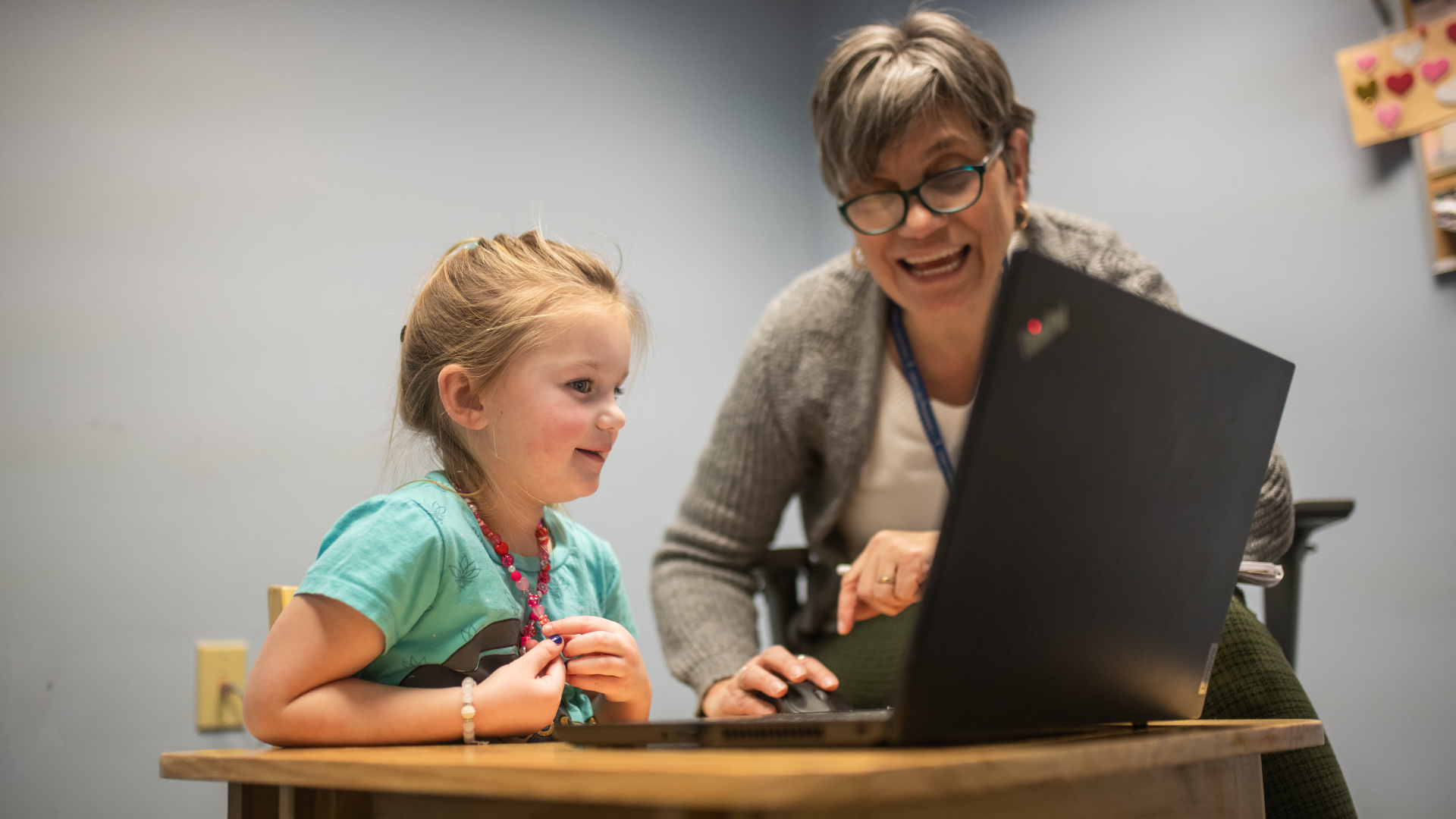 A child in a light blue shirt at a table looking at a computer with a therapist.