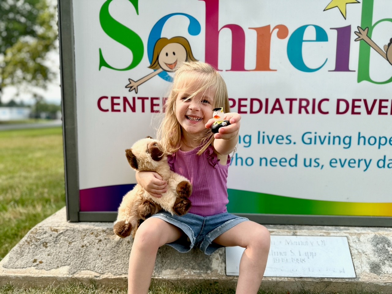 A child sitting and smiling in front of a Schreiber Center of Pediatric Development sign holding a stuffed animial.