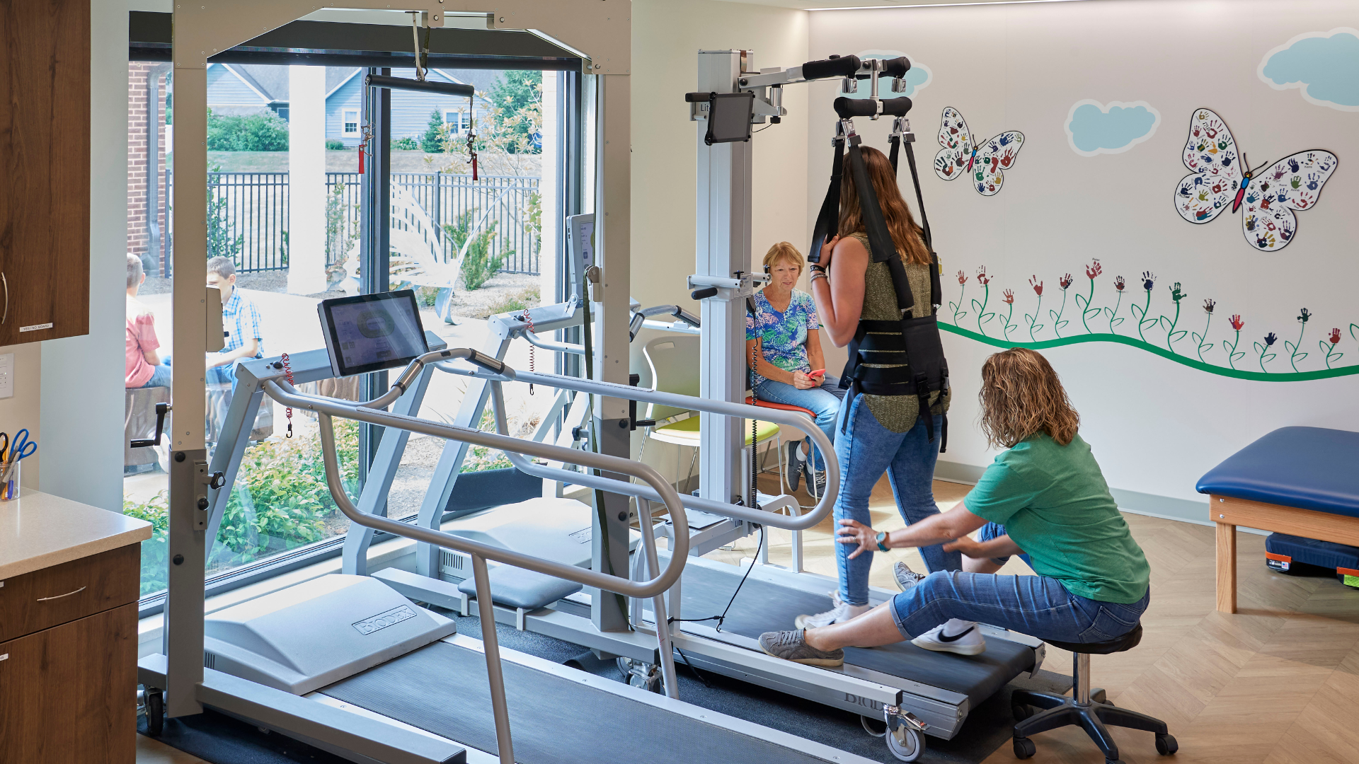 A person utilizing an overhead track harness to walk on a treadmill with an adult guiding their legs.